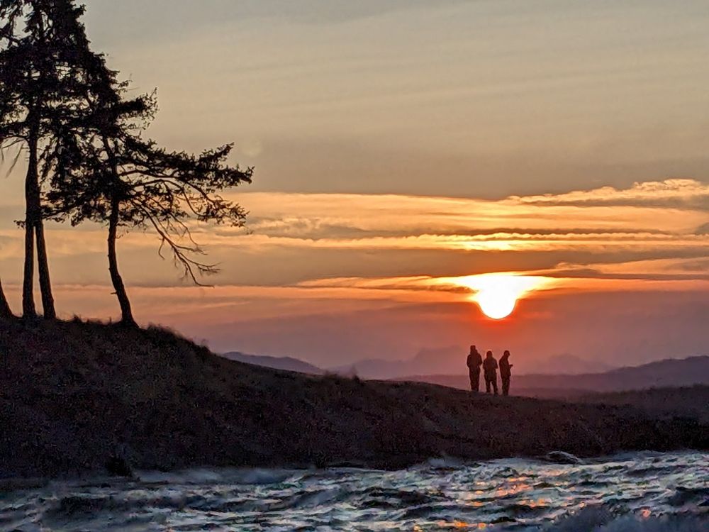 A rocky promontory with three people standing silhouetted under the setting sun. Three big Douglas firs off to the side create some photographic alliteration. 
