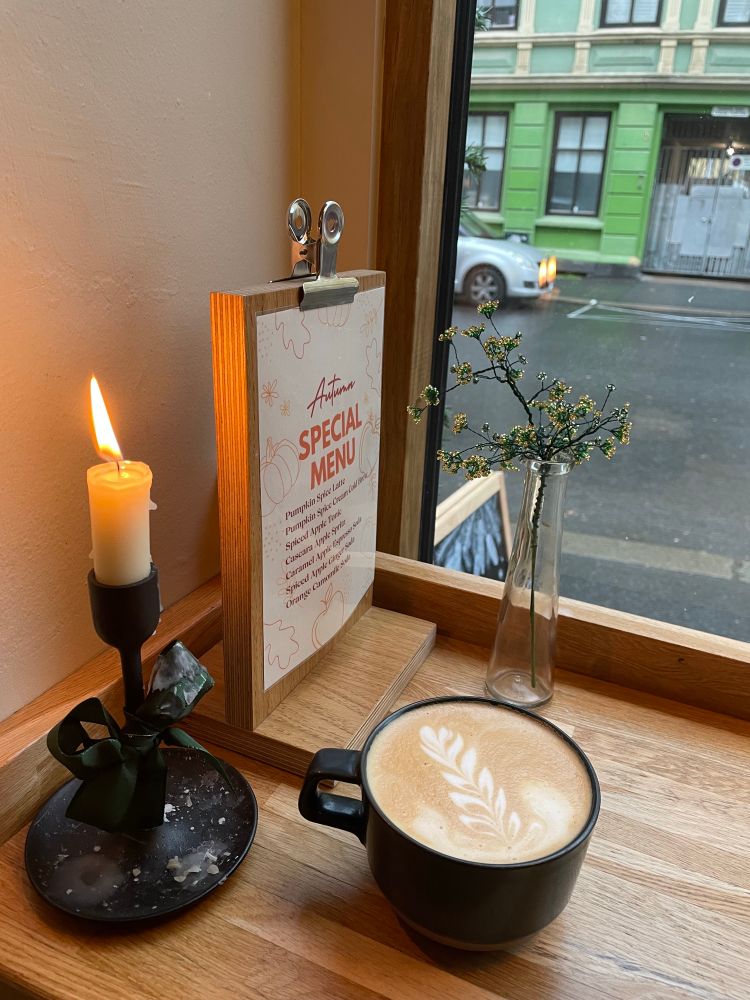 A little latte vignette with a lot candle, cup of coffee with leaf latte art, a little sign saying the autumn specials, and a little vase of beaded Xmas decor in a cafe window in Oslo overlooking a rainy street. 