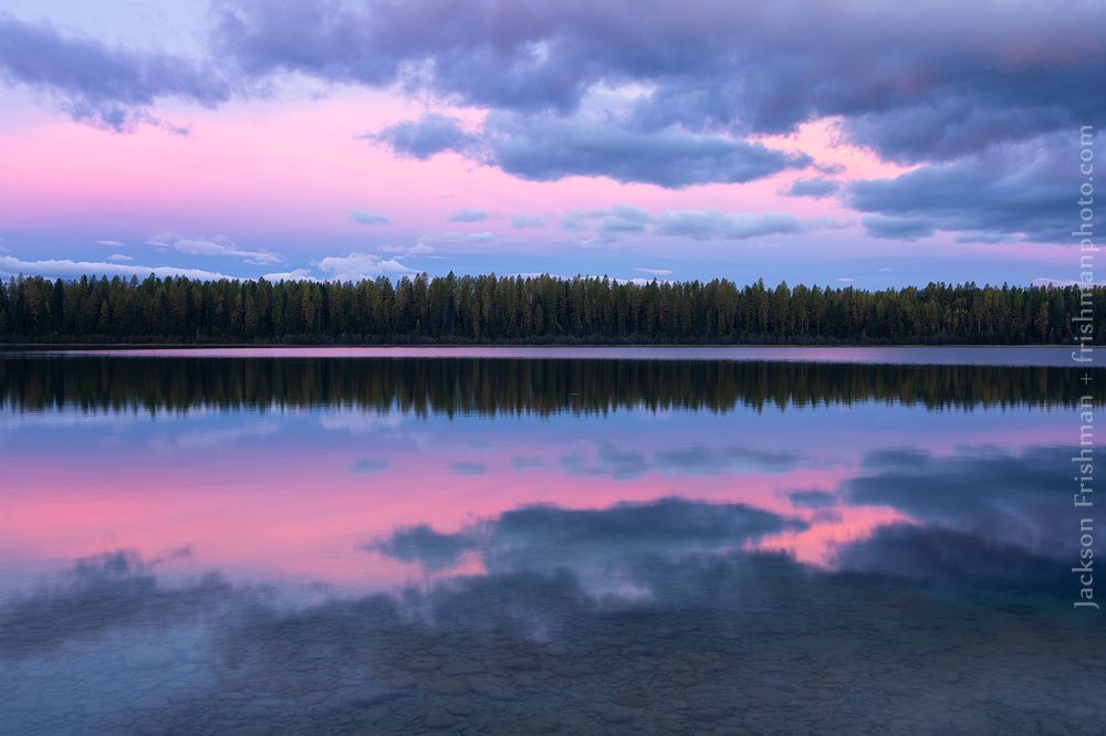 Photograph of early sunrise reflected in a still lake. The far shore of the lake is forested with conifers. Blue clouds showing just a hint of purple and their reflections form an oval frame around woods and lake. The sky behind the clouds is glowing with pre-dawn color of blue and purple grading into subtle orange, colors which are reflected richly in the calm water. 