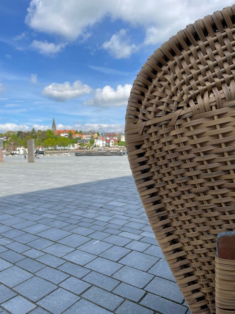 Blick an einem Strandkorb vorbei über eine Uferpromenade. Im Hintergrund sind Häuser und ein Kirchturm zu sehen mit einigen Bäumen. Der Himmel ist blau mit einigen Wolken