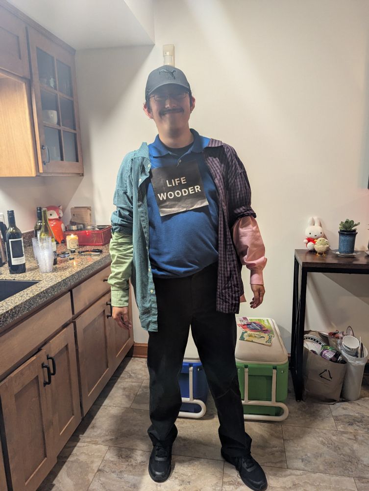 Me dressed in greens, blues, purples, and pinks with a black cap on. I'm wearing a label that says "Life Wooder".