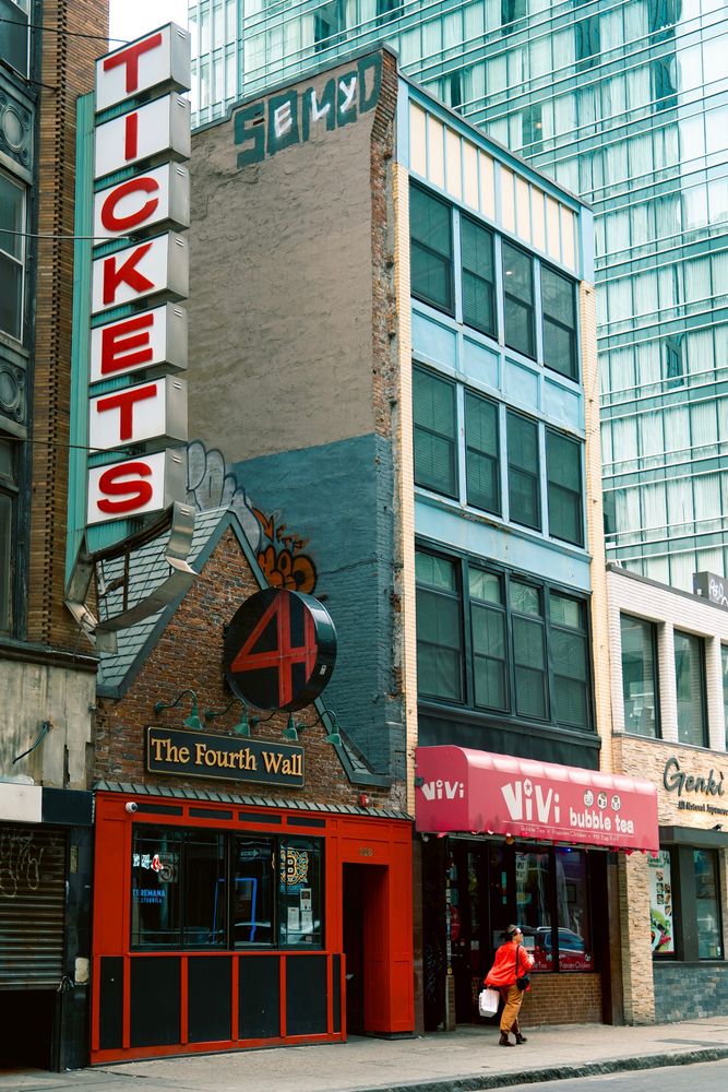 A woman in red walking past buildings in Chinatown, Boston. A retro-style vertical tickets sign featured prominently in the top left of the composition.