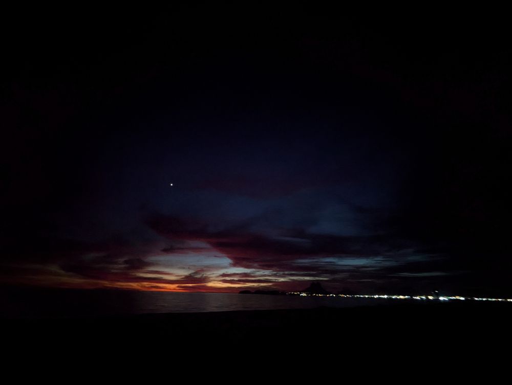 Mostly dark sky over the Sea of Cortez as the last sunset hues slip away in deep red and orange. Mars is clearly visible and lights illuminate the coastline. 