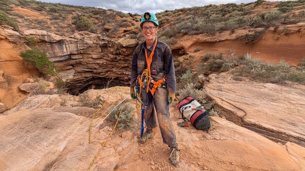 A woman (me!!) in dusty overalls standing next to the entrance of a cave, wearing a green helmet, a harness and gear for rope work. 
