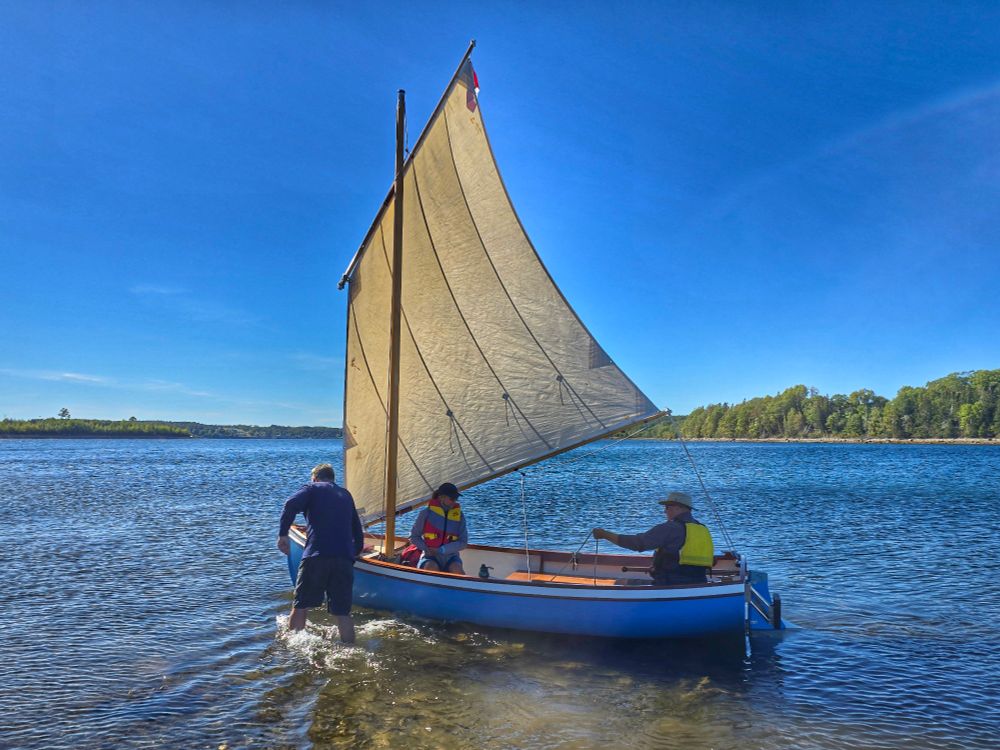 A man pushes a small balanced lug sailboat off a beach with a man and woman aboard.  