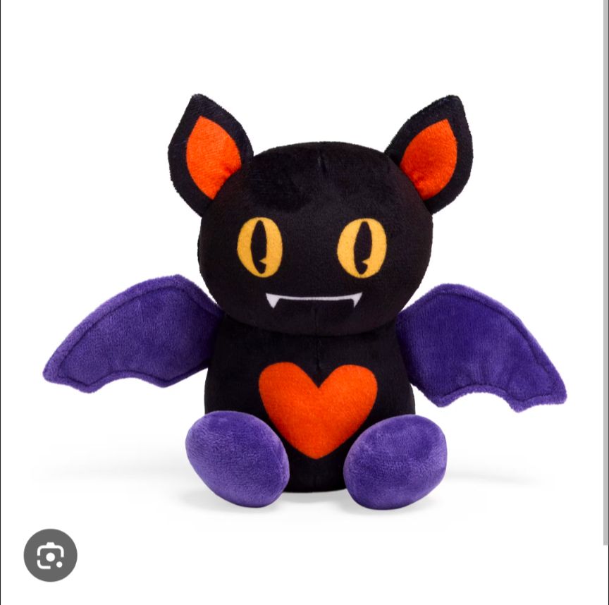 A black stuffed bat with big yellow eyes, tiny vampire fangs, a red heart tummy, and purple wings and feet.