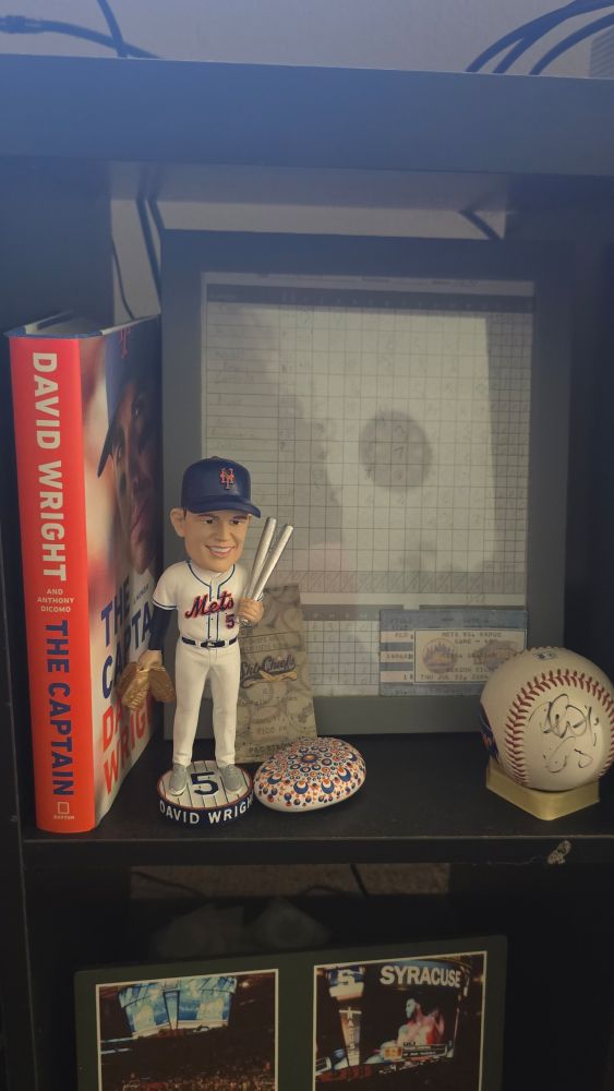 Shelf containing David Wright's book "The Captain," his bobblehead, ticket stubs, an autographed ball, a signed baseball, and a rock painted blue and orange. 