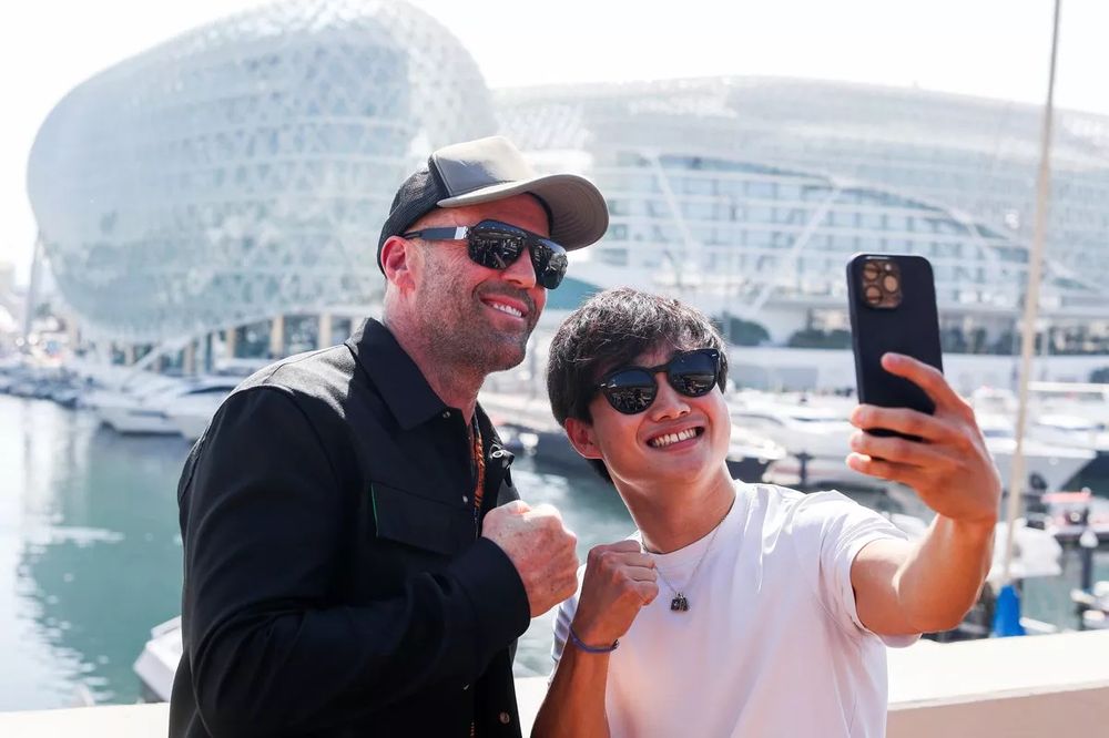Jason Statham and Yuki Tsunoda with the hotel on the track in Abu Dhabi in the background each making a fist and smiling while Yuki holds his phone up to take a picture