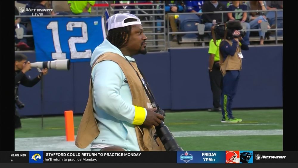 Marshawn in a backwards hat, seafoam hoodie, tan photographer pullover holding a long lens camera on a strap around his neck