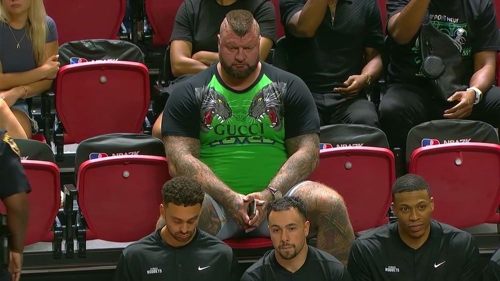 Wider shot of sizeable man with a beard and pointed mohawk in a green Gucci shirt with black short sleeves that has panthers on both shoulders - think Zangief