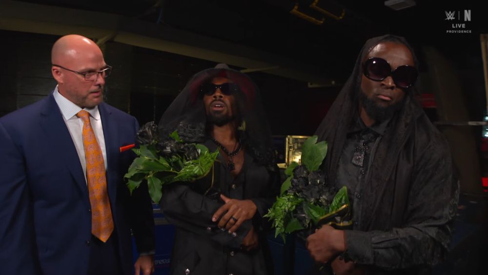 Adam Pearce (WWE) in a blue suit with an orange tie looking at Xavier Woods and Kofie Kingston in all black, sunglasses, and veils, with black flowers 