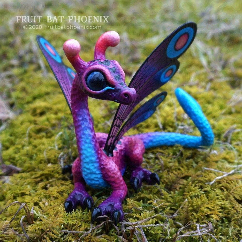 A small, cute, big-eyed blue, purple, and pink dragon sculpture with butterfly-looking wings.