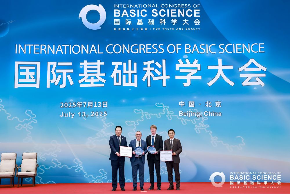 Four scientists stand in suits, holding certificates and awards, in front of a large blue screen for the International Congress of Basic Science