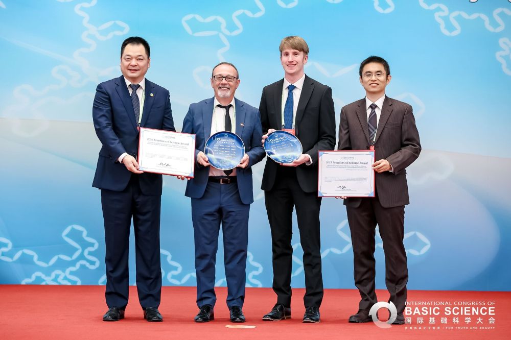 Four scientists stand in suits, holding certificates and awards, in front of a large blue screen for the International Congress of Basic Science