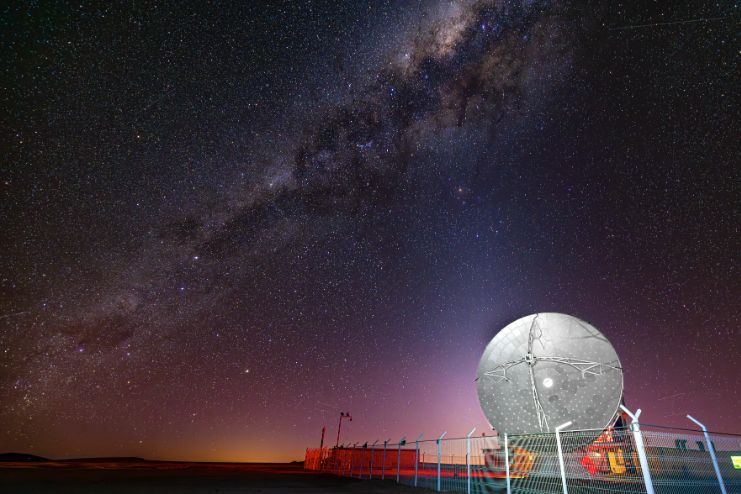 The APEX telescope sits in front of an orange glow of sunrise and beneath the starry sky and Milky Way