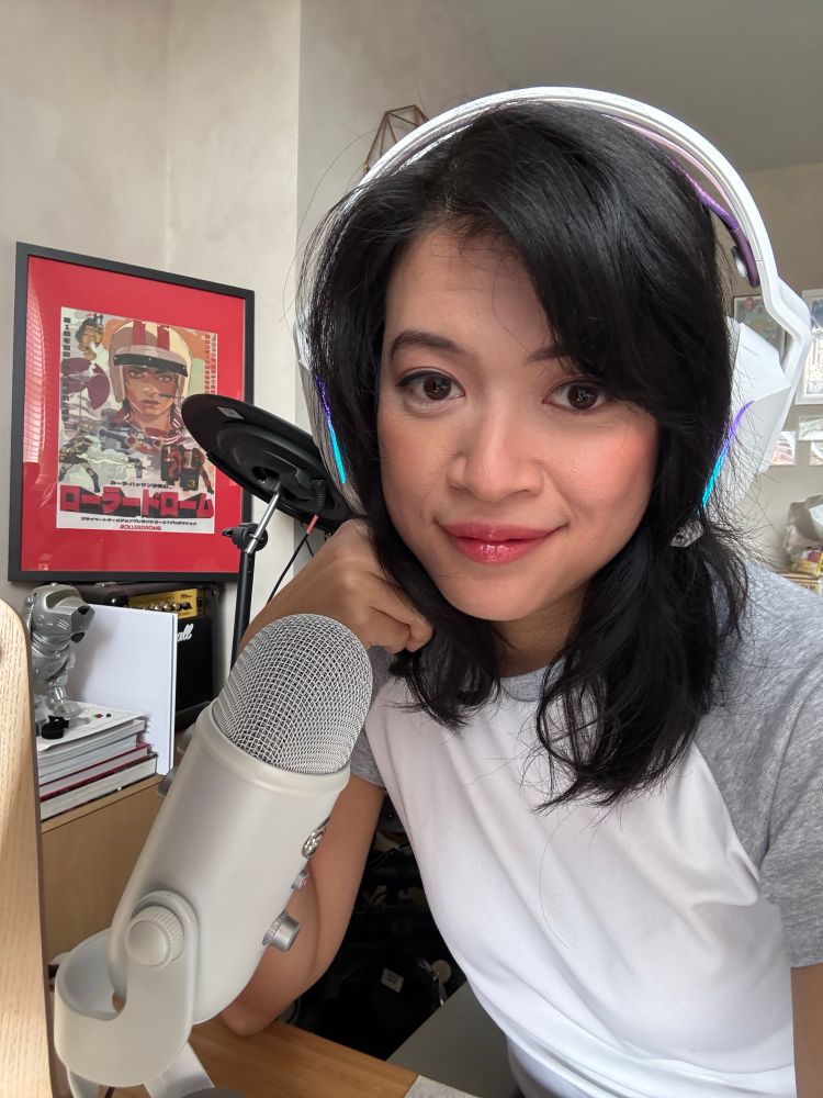 Anisa at her work desk wearing headphones in front of a mic. A poster of rollerdrome can be seen in the background. 