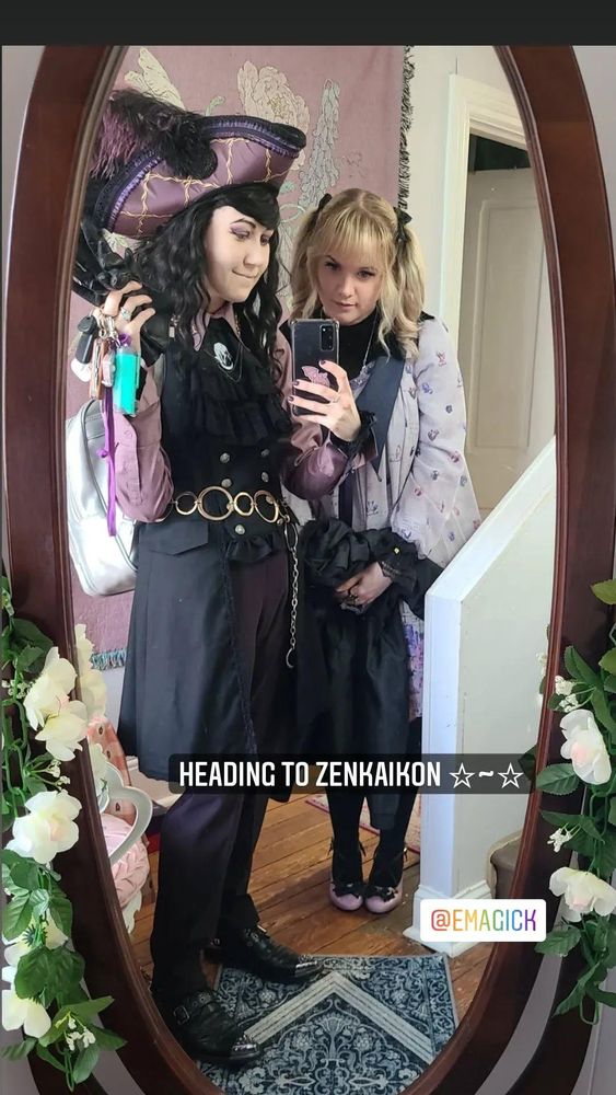 Emi and Mar are standing in front of a standing mirrror. text on sceen announces theylir departureto zenkaikon. Emi is wearing Magical Stone OP by Metamorphose with her hair in pink tails. Accompanied with a starcrossedlovies moth ring and a lavender cotton candy fantasy bag. Mar is wearing a Scarlett Rose tricorn pirate hat, with a lavender blouse, Alice and the Pirates gilet, with a f+f cravat. Their cravat is pinned with a coey Unicorn enamel pin, along with a neate glass coffin ring. both individuals wearing a mix of black and cool toned lavender.