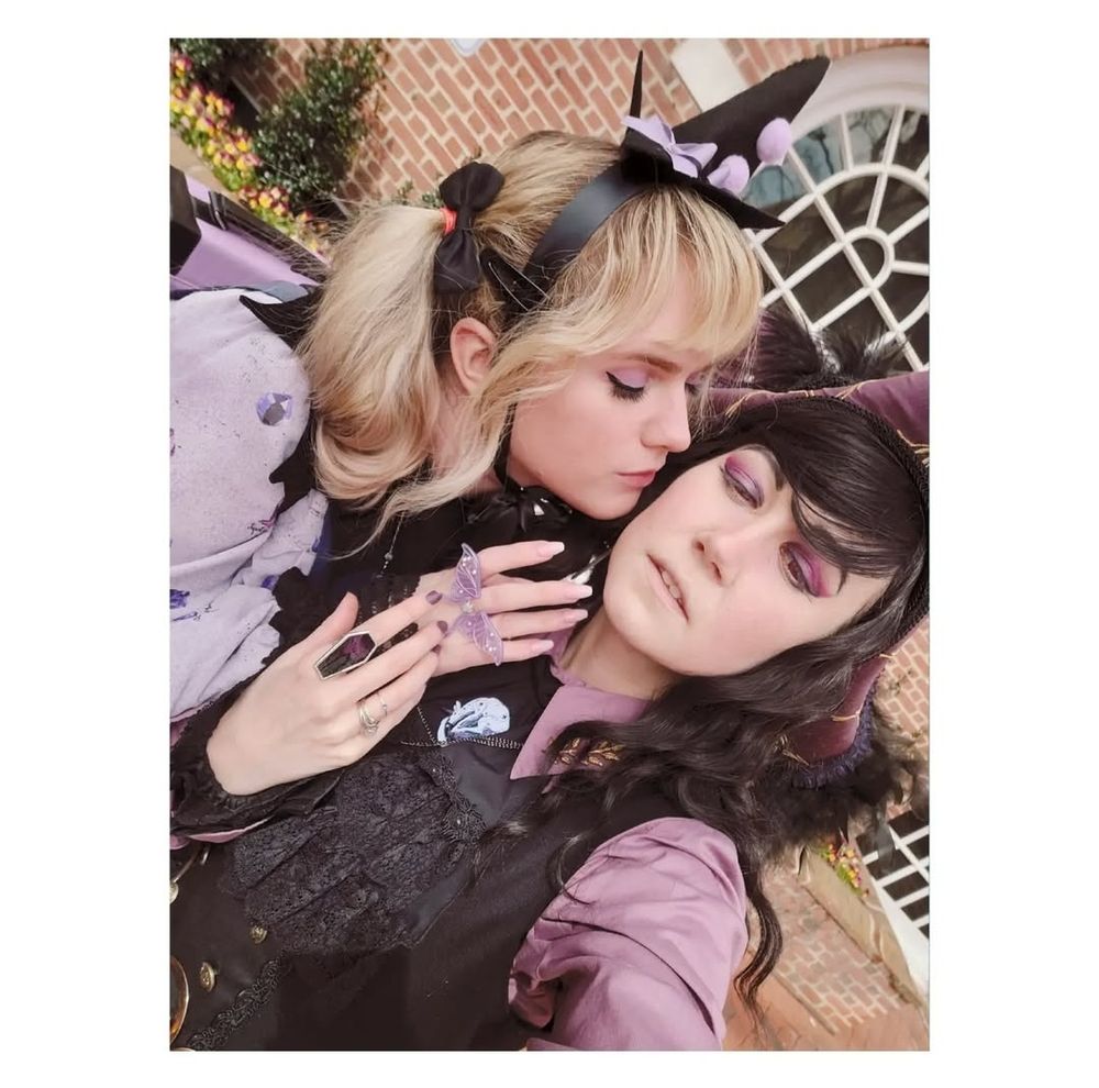 Emi kissing Mar lightly on the cheek as touch hands. Emi is wearing Magical Stone OP by Metamorphose with a handmade tiny witch hat. Accompanied with a starcrossedlovies moth ring and a lavender cotton candy fantasy bag. Mar is wearing a Scarlett Rose tricorn pirate hat, with a lavender blouse, Alice and the Pirates gilet, with a f+f cravat. Their cravat is pinned with a coey Unicorn enamel pin, along with a neate glass coffin ring. both individuals wearing a mix of black and cool toned lavender.