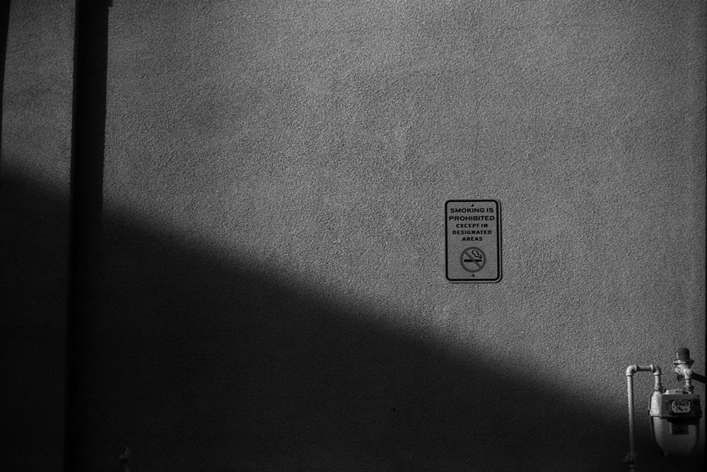 I black and white film photograph of a wall with a 'No Smoking' sign posted. A dark shadow crosses the frame diagonally in the lower left-hand corner.