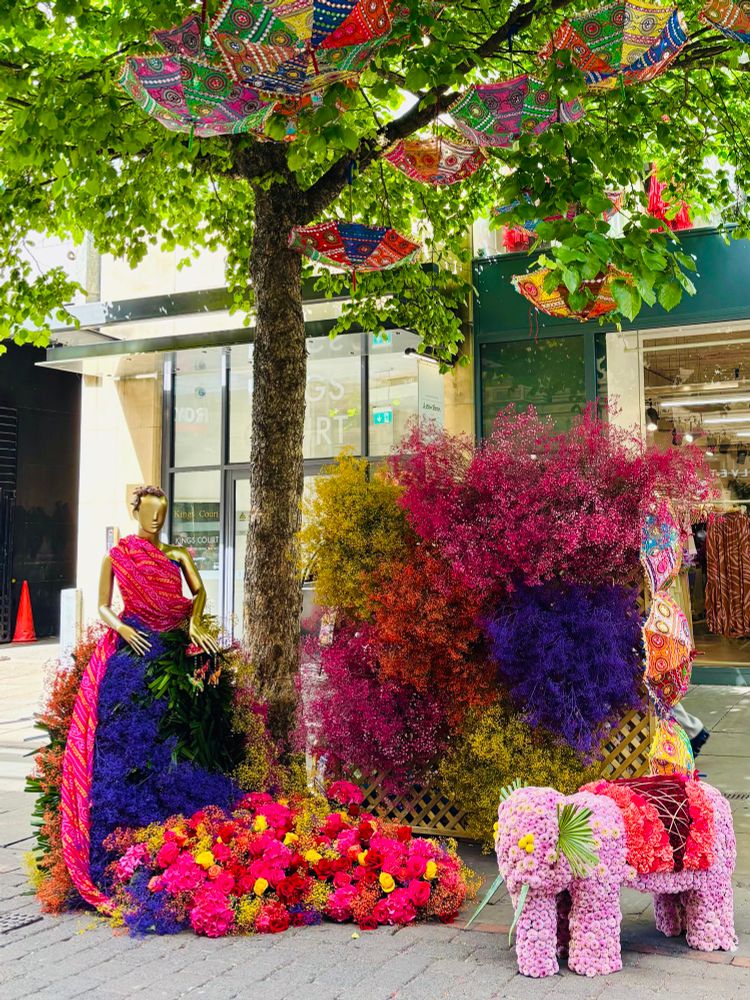 A vibrant floral installation beneath a leafy tree, featuring a mannequin dressed in a skirt made of purple and green flowers, with a bright pink and gold sash. Colourful parasols hang overhead, and a small pink elephant sculpture made entirely of flowers sits nearby.