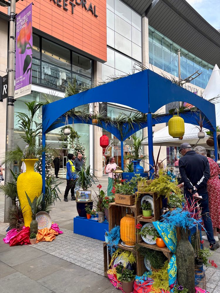 A street scene from the Manchester Flower Festival on New Cathedral Street. A bright blue structure frames an arrangement of tropical plants, bold fabric drapes and painted crates filled with flowers, oranges and decorative plates. Festival banners and people browsing the displays complete the scene. 
