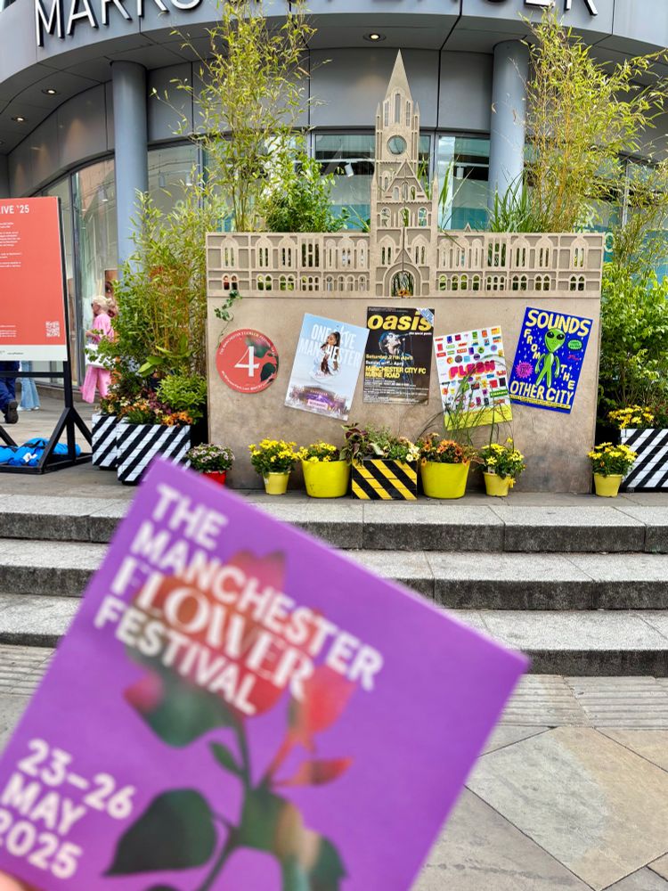 A close-up of a Manchester Flower Festival flyer in the foreground, held up in front of a floral display shaped like Manchester Town Hall. Behind it are posters referencing Manchester culture (Oasis, Sounds from the Other City) and colourful potted plants arranged at the base of the structure.