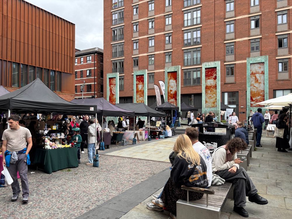 Stalls and shoppers at the Ancoats Makers Market, set against red-brick apartment buildings, with people browsing handmade goods and sitting on benches.