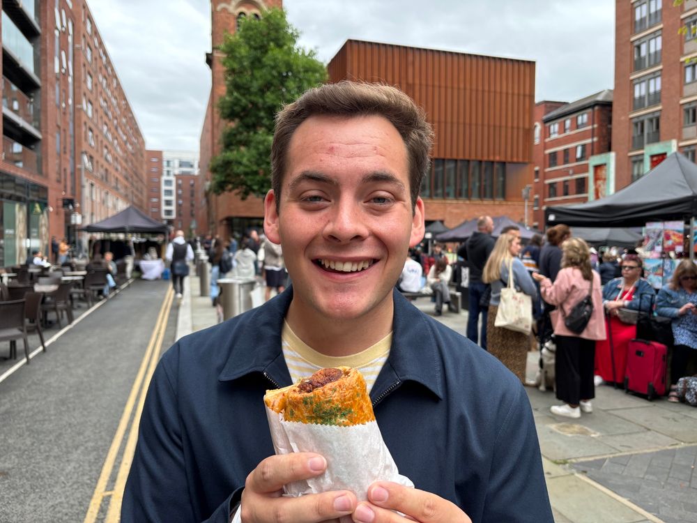 A smiling Luke holds a pastry wrapped in white paper at the Ancoats Makers Market, with red-brick buildings and market stalls visible in the background.