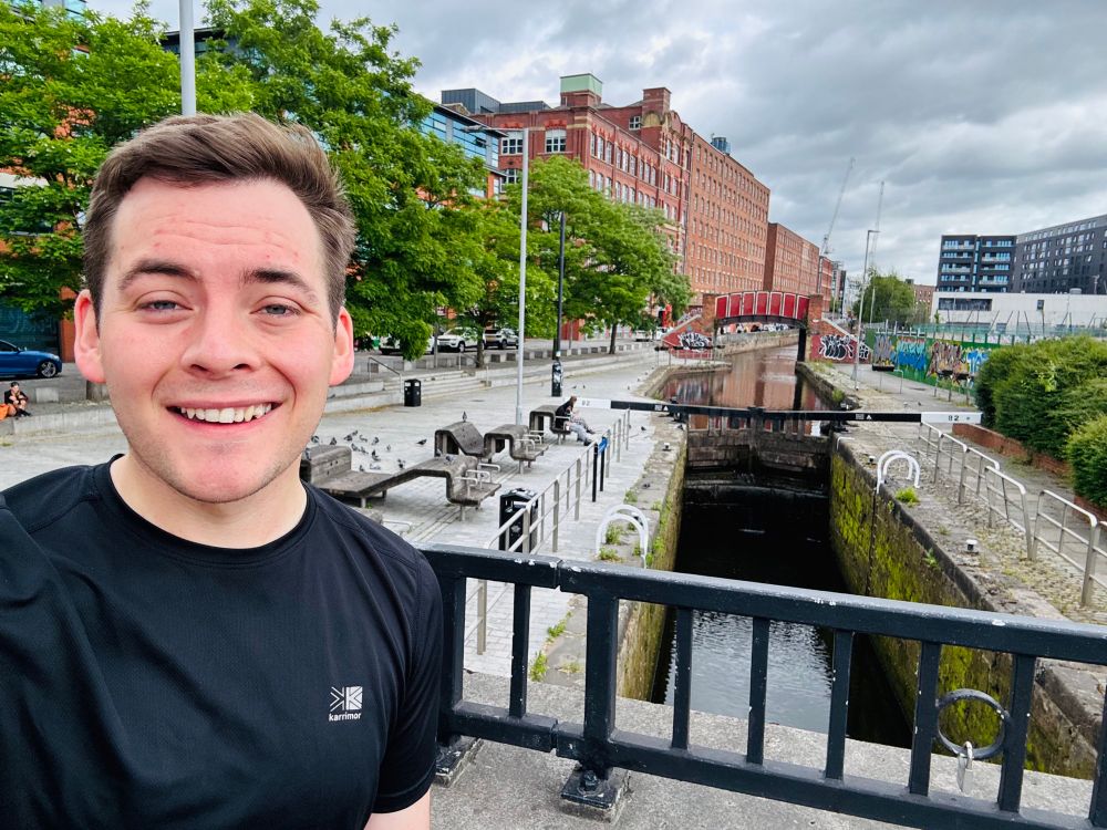 Luke, smiling in gym gear, stands in front of a canal lock with red-brick buildings, trees and street art in the background. It’s a cloudy bank holiday, but the weather looks dry.
