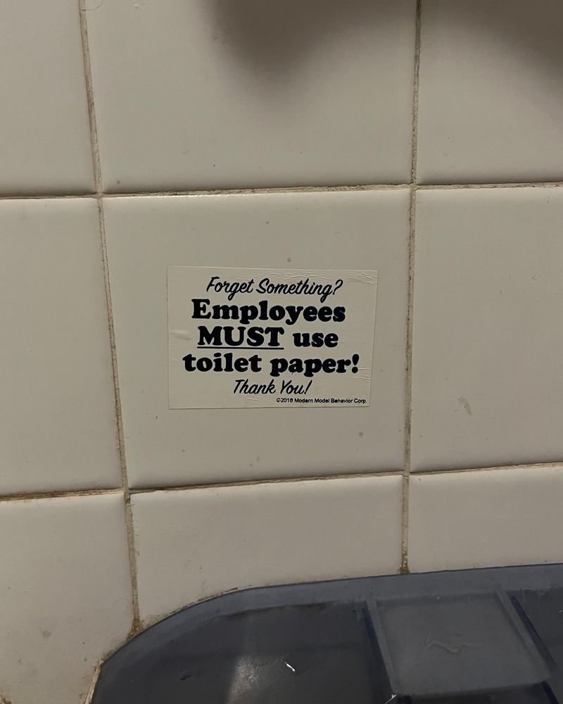 Grimy tile wall in a bathroom. A sticker above the toilet paper dispenser reads:

Forget Something?
Employees MUST use toilet paper!
Thank You!