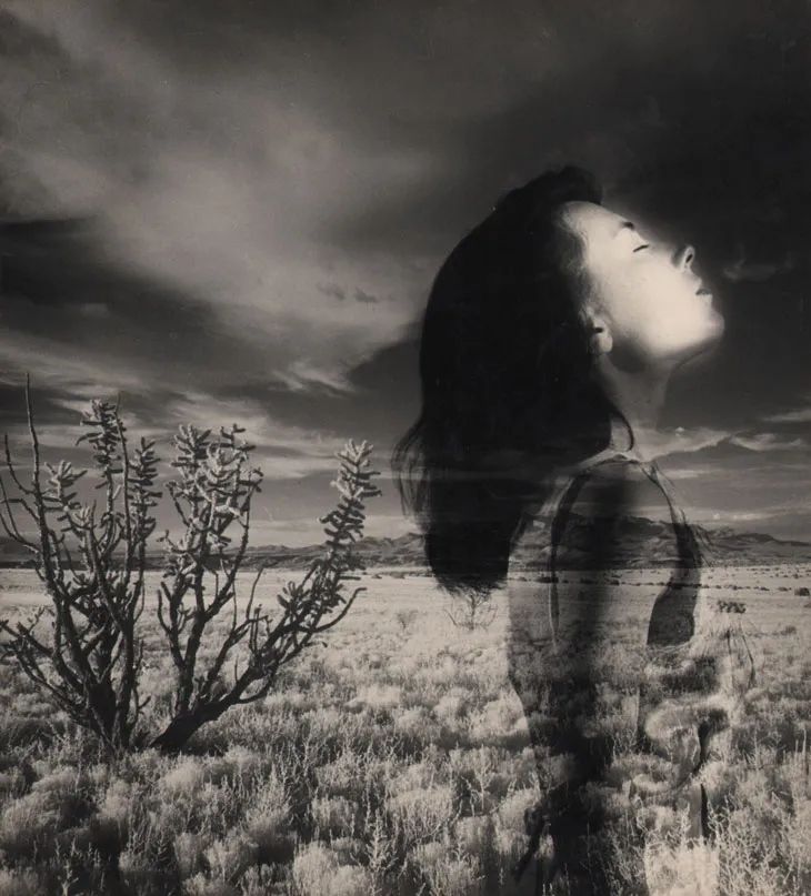 This ethereal black-and-white image is a double exposure photograph combining a serene desert landscape with the profile of a woman. Her face is tilted upward, eyes closed, as if basking in sunlight or lost in a moment of peaceful introspection. The arid terrain, dotted with sparse shrubs and silhouetted mountains in the distance, merges seamlessly with her translucent figure, creating a haunting fusion of human and nature.