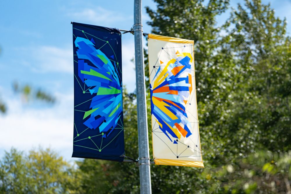 Two banners erected on a streetlight poll. On each banner is one side of a butterfly in abstract, fragmented form with one banner in shades of blues and green and the other in shades of yellows, blues and orange.