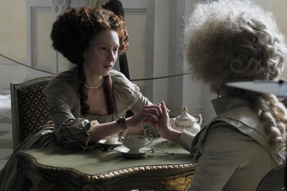 Roxane Duran in the role of Princess Lamballe. She’s seating at the table with Queen Marie Antoinette and holding her hand. They seem to be very good friends.