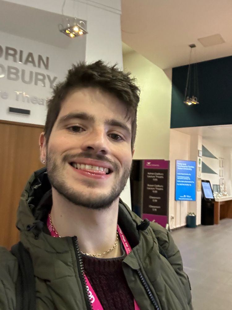 David taking a selfie in the lobby of a hotel in a red jumper, green coat and pink lanyard. 