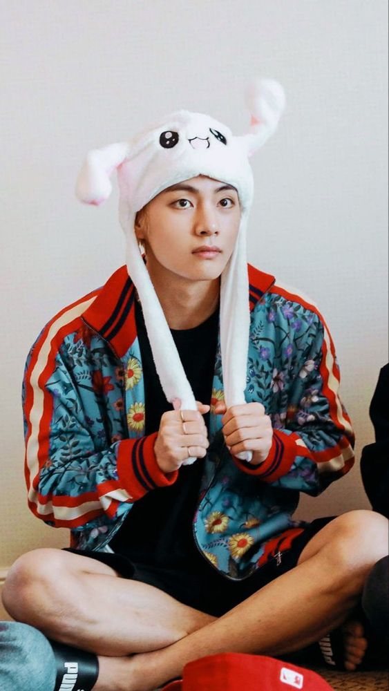 Tae in black shorts, puma sliders, a rabbit hat, black shirt and very colorful jack, red and black and flowers.  He looks serious even though he is being silly.