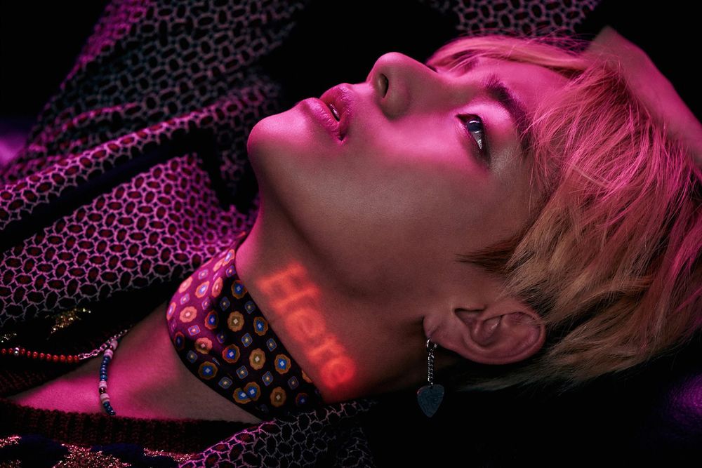 Taehyung laying back, blond hair, multi color ahem color around his neck, the word Here glowing on his neck. He is wearing an earring that sure looks like Yoongi's future D-Day guitar pic but of course it isn't, probably. Never know with them.  They are crafty.  He has on a very patterned jacket, over a black and gold vest(?).  He is looking up at the ceiling, and a pink light glows over his face and body.  Delicious.