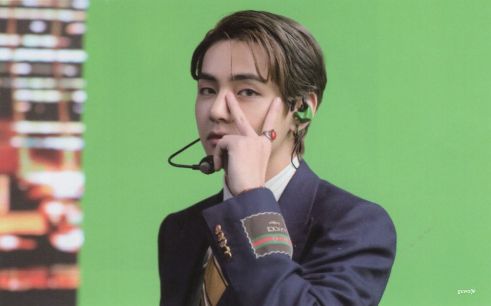 Slick haired Tae doing his V sign, in front of green screen thing, wearing blazer and white shirt and tie.  