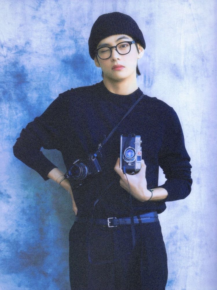 Tae winking at the camera. He’s holding his own camera, wearing all black, including a black beanie and black framed glasses.