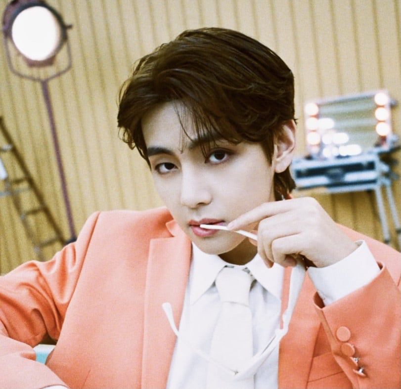 Butter era Tae in a salmon orange suit, holding a pair of sunglasses, the arm pressed to his lips. 