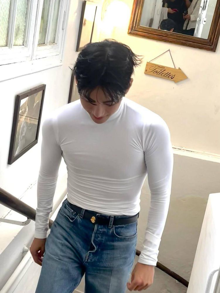 Tae in a very fitted white long sleeve shirt and jeans. He’s walking up stairs, facing the camera and looking down with what looks like a faint smile. 