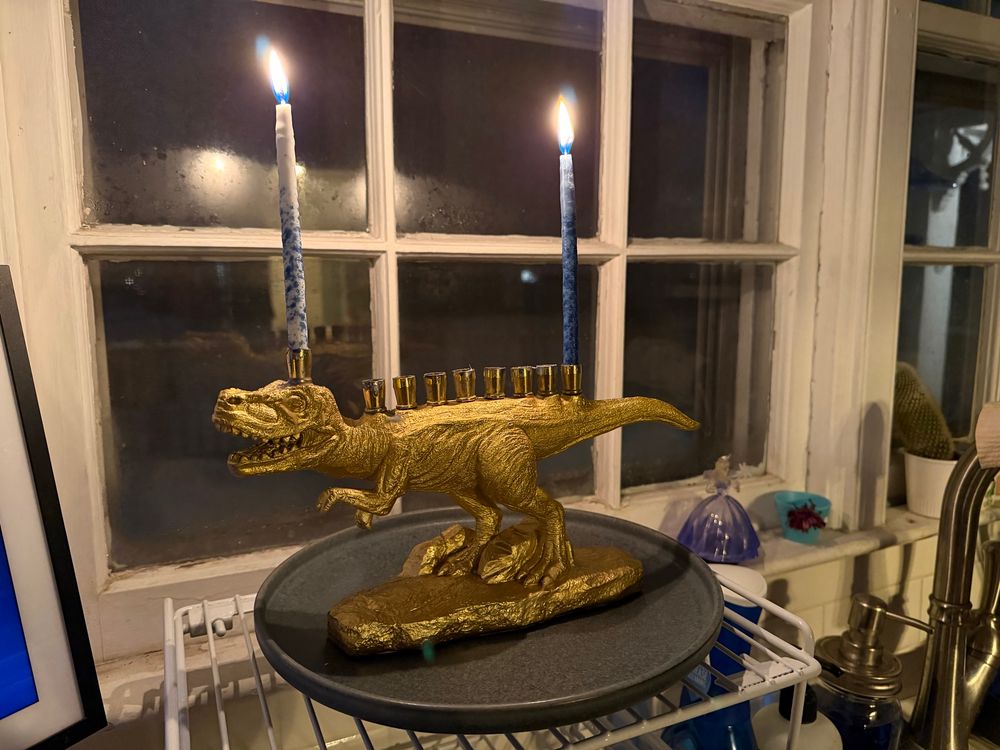 A gold-guilded menorah in the shape of a Tyrannosaurus Rex sits on a plate in front of a frosted kitchen window. The shamash candle is lit atop its head, and the candle for the first night of Hanukkah is lit at the base of its tail.