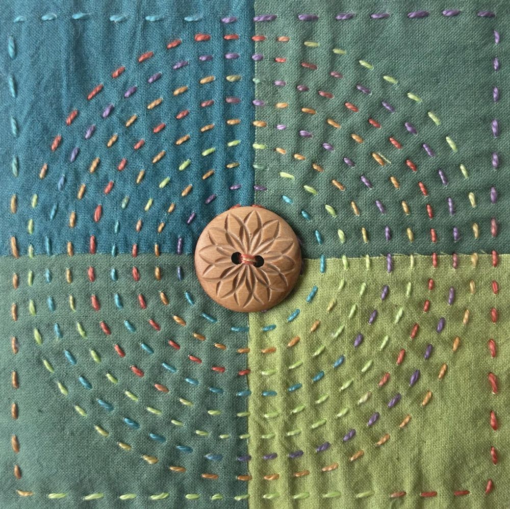 A 4x4 inch square made with bright coloured cotton fabric scraps, in the centre a wooden button. Stitched with variegated thread in circles. Stop killing the children!