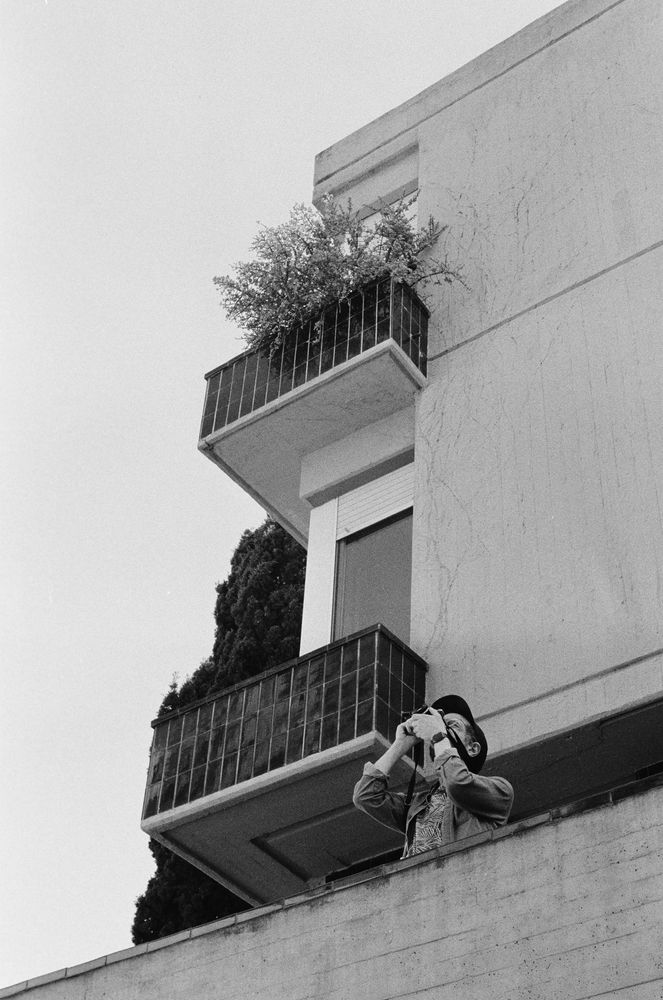 Black and white photograph of a man taking a photo from a balcony. The photograph was taken from a lower floor; you can see a building and a tree behind him.