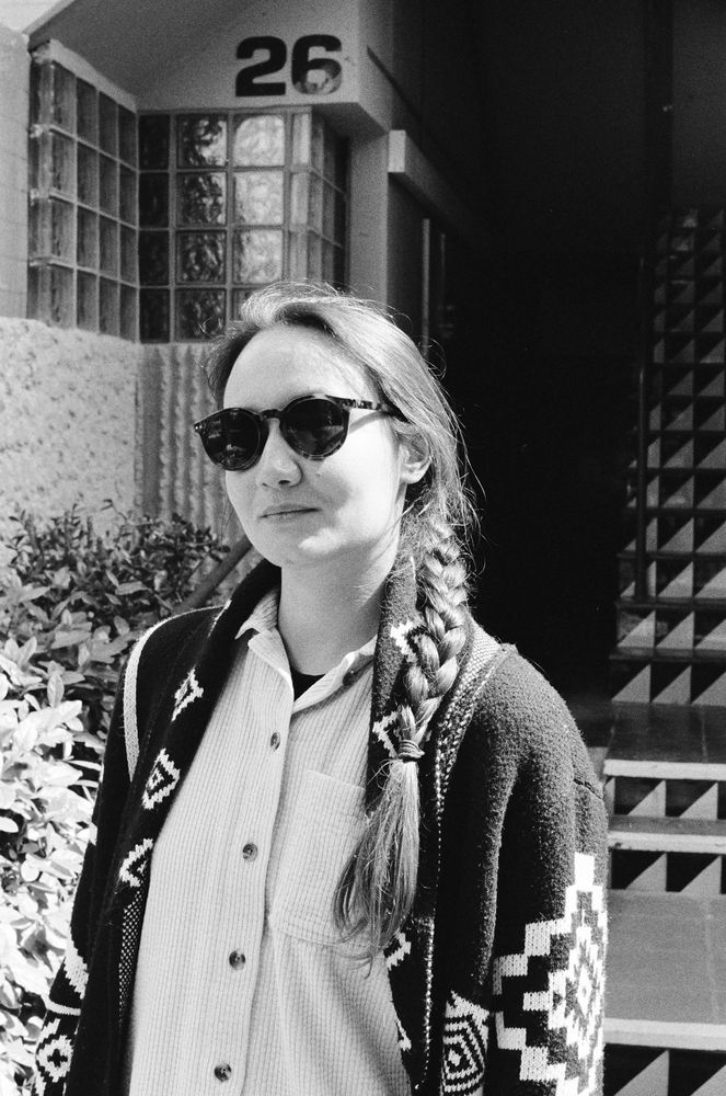 Black and white close-up portrait of a woman wearing sunglasses, a white shirt, and a patterned jacket, standing in front of a building. Her hair is braided and falls over her shoulder.