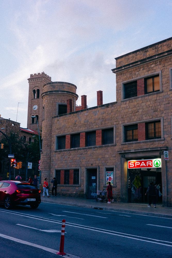 Fotografía al atardecer de un supermercado Spar en un edificio de piedra al que solo le falta un foso con cocodrilos. Frente al edificio se ve gente y coches circulando.

A sunset photograph of a Spar supermarket in a stone building that only needs a moat with crocodiles. People and cars can be seen driving in front of the building.