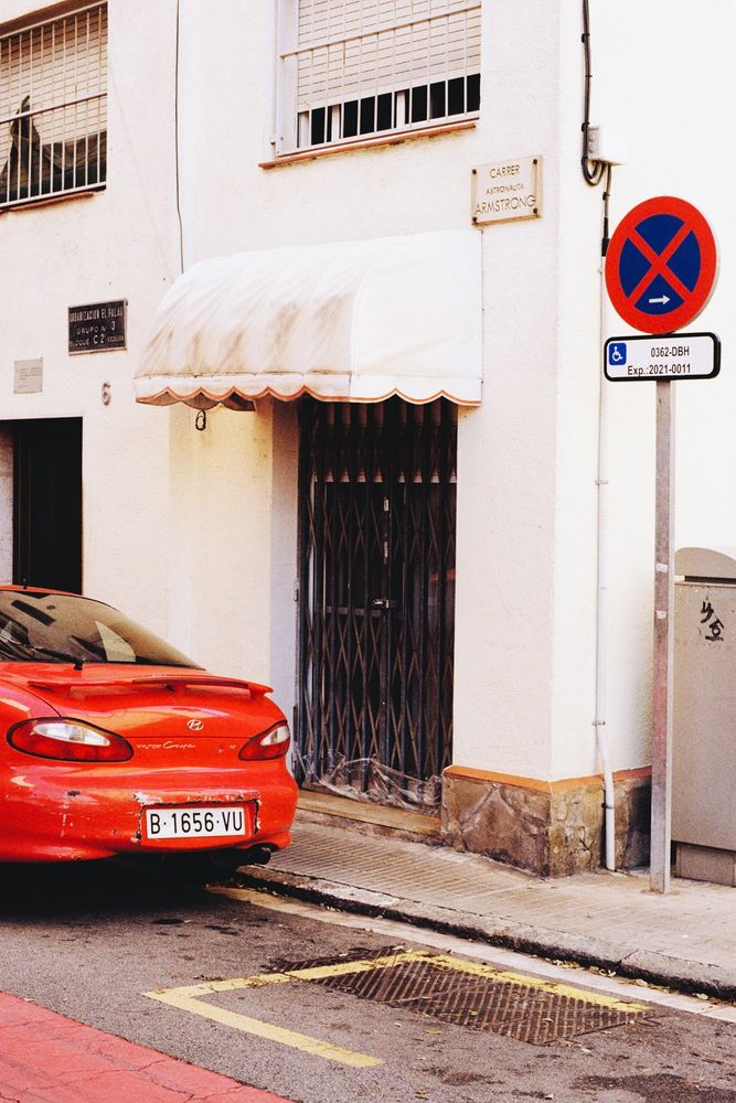 Photograph of a building door with bars and a white awning. In front of it there is a red car parked, a no parking sign and a section of asphalt painted red.