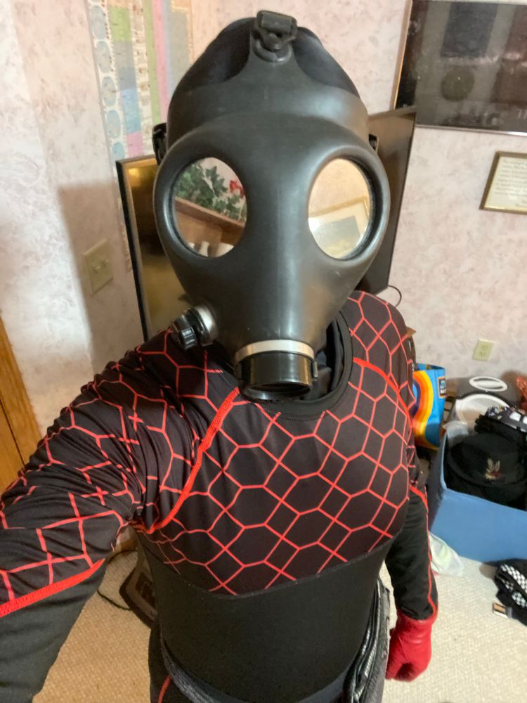 W84 stands in a bedroom in front of a tv, in its full drone outfit. Its leggings and long sleeve shirt are both black with a red hex pattern, it’s wearing red gloves, a black balaclava tucked into its shirt, and it’s Israeli style gas mask with mirrored lenses