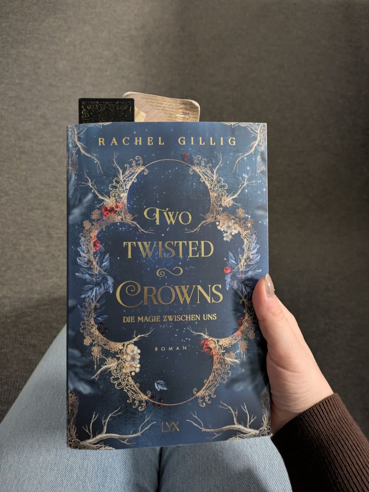 Auf dem Foto sieht man, wie ich das Buch Two Twisted Crowns - Die Magie zwischen uns von Rachel Gillig in der Hand halte. Das Buchcover ist dunkel gehalten. Die Schrift des Titels ist verschnörkelt und golden. Umrahmt wird der Titel von einer goldenen Umrandung, die aus Ästen und bunten Blumen besteht.