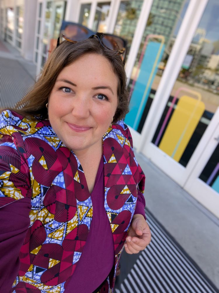 Selfie of smiling woman in pink.sjort and graphic block print jacket in front of windows with stylized bar chart components.