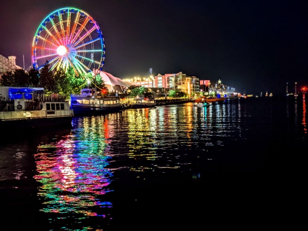The lights at the Chicago Navy Pier shine bright at night. It's the summer solstice and visitors, as well as locals, enjoy a night out in town. The multicolored lights of the Ferris Wheel and other attractions reflect on the waters of the lake. A Water Taxi is docked on the pier. 
